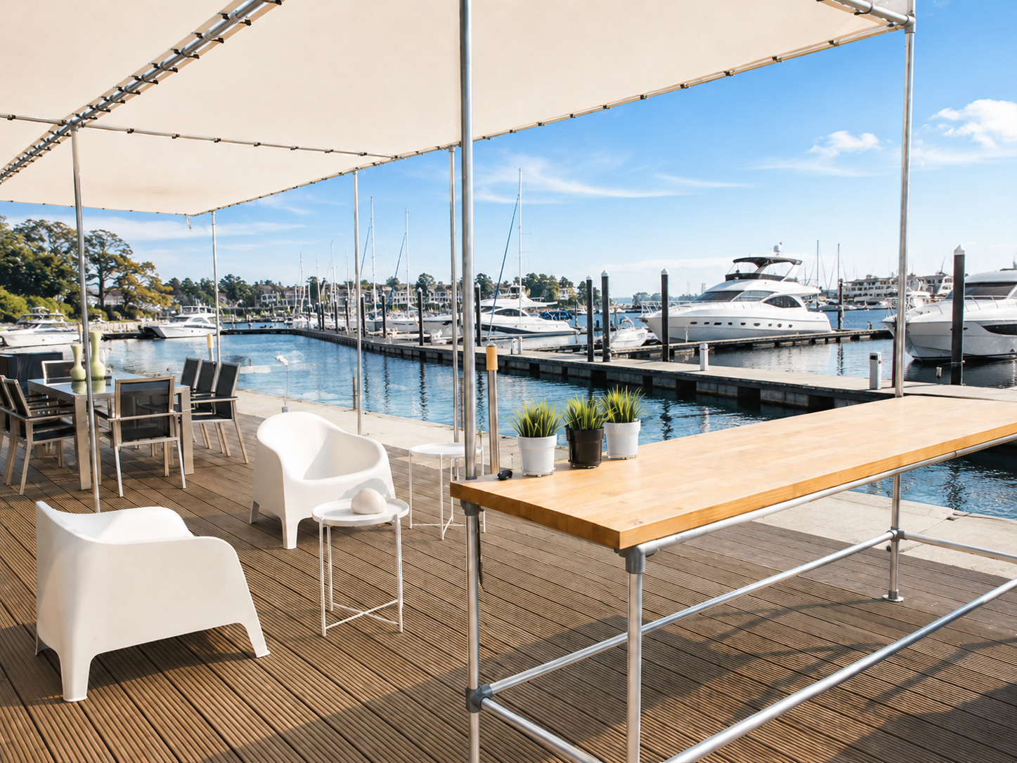 Terraza de madera bajo un toldo en el puerto deportivo. Modernos sillones blancos de salón, mesa de comedor y una larga barra de madera con plantas en macetas. Al fondo, numerosos veleros y yates en las aguas azules del puerto bajo un cielo azul radiante.