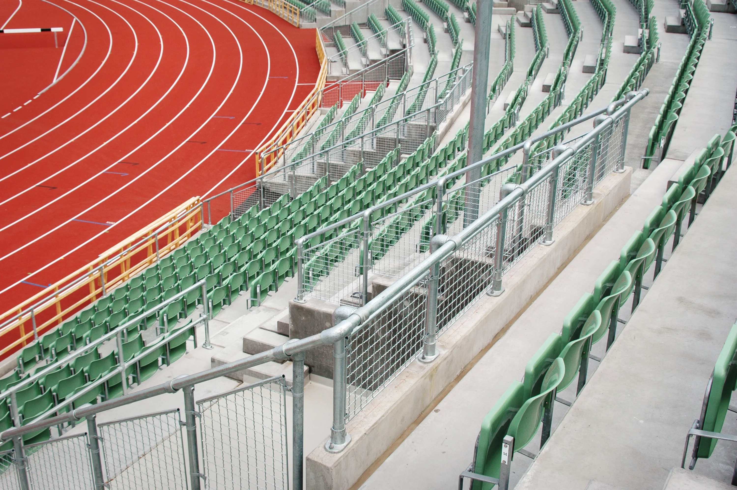 An empty red running track with white lane markings is on the left and curves to the right. On the right and in the background are empty green stadium seats on concrete steps, separated by metal railings with mesh.