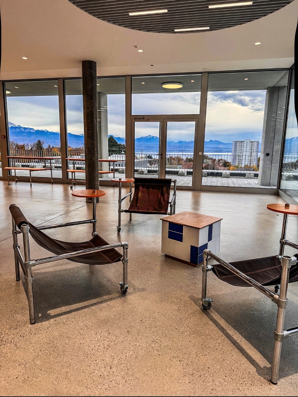 A modern lounge area with large windows and sweeping views of the lake, snow-capped mountains and a cloudy sky. Three brown leather armchairs with chrome frames on castors, along with small orange tables, stand on a light-coloured terrazzo floor.