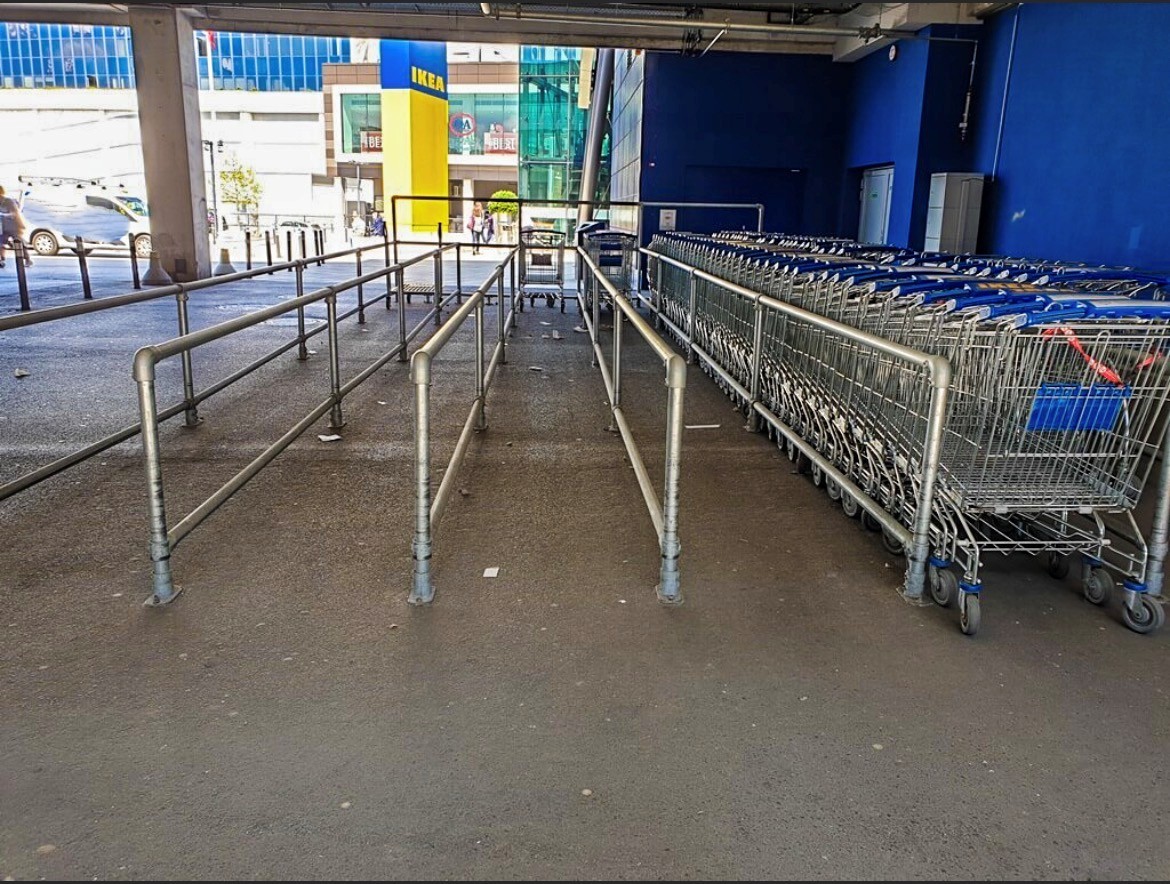 Zona de acceso a una tienda IKEA: a la derecha, una larga fila de carros de compra apilados con asas azules. A la izquierda, barreras metálicas guían al cliente por el suelo gris hasta la entrada. Al fondo, el logotipo de IKEA.