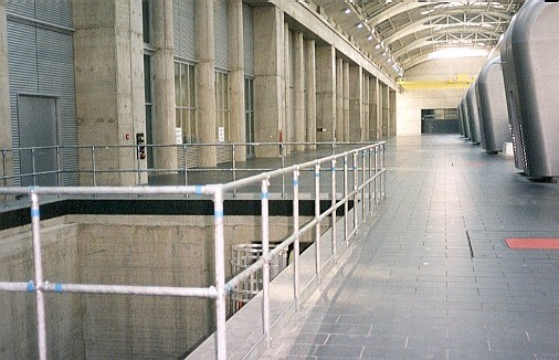 Nave industrial amplia y luminosa, paredes de hormigón y ventanas altas. Puerta enrollable a la izquierda, barandilla ante hueco de ascensor. Suelo de baldosas oscuras, máquinas a la derecha y grúa amarilla en el techo.