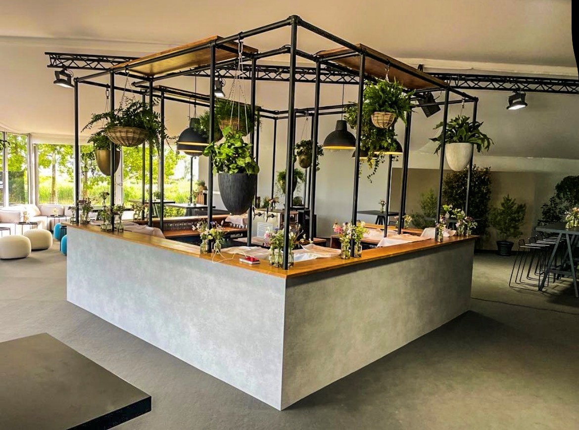 Barra moderna de hormigón gris y encimera de madera en una estancia luminosa. Arriba, estructura metálica negra con plantas y lámparas colgantes. La barra tiene pequeños arreglos florales. Ventanales a un paisaje verde.