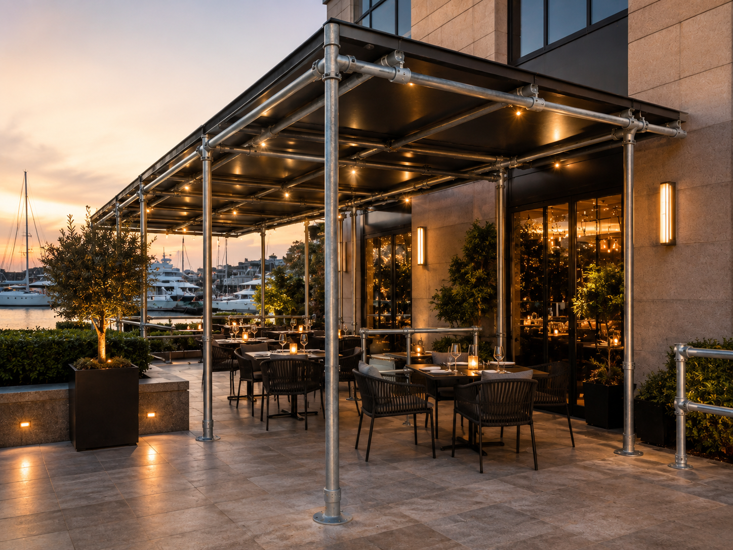 Terraza de un restaurante en el puerto deportivo al atardecer. Cubierta decorada con guirnaldas de luces, mesas con copas y velas. Barcos al fondo, cielo de tonos naranja y violeta, plantas y luces de suelo.